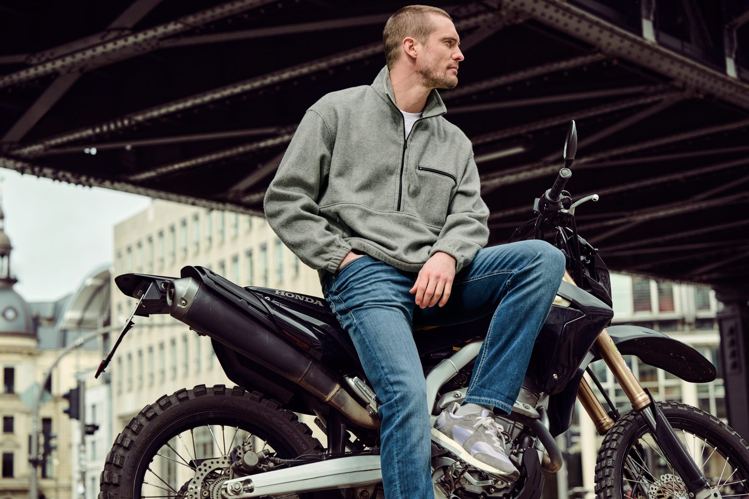 Man wearing NEFLYX jeans seated on a motorcycle beneath a steel bridge