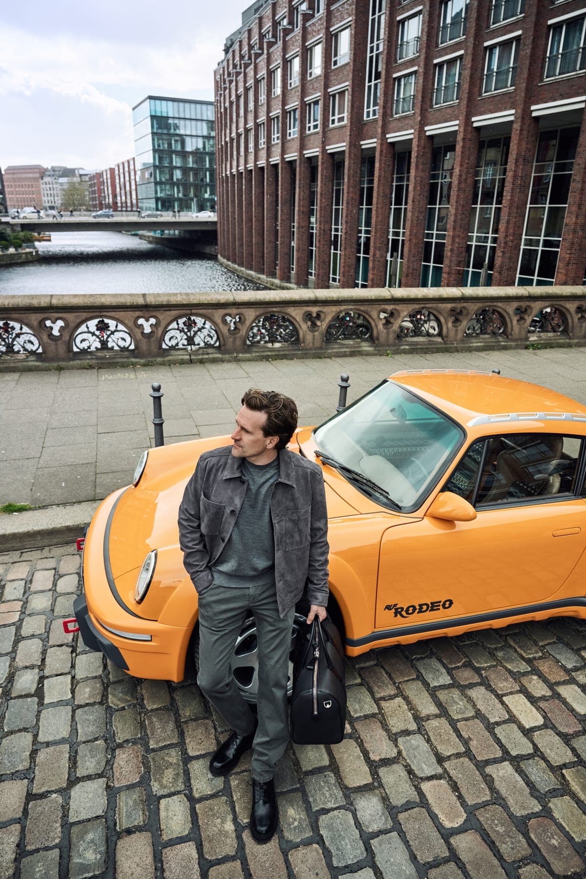 Man in NEFLYX denim leaning against an orange sports car by the harbour
